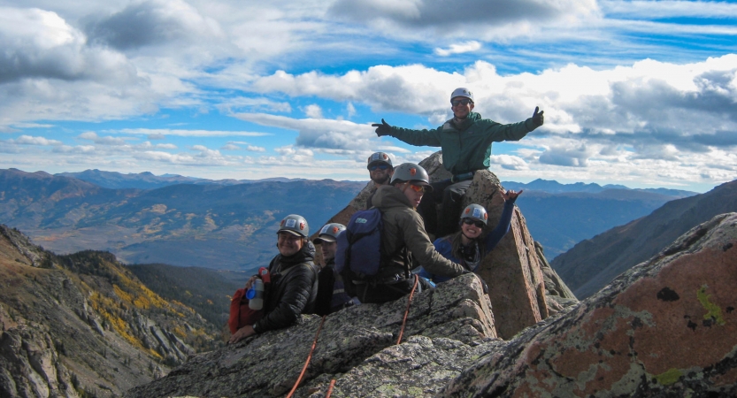 A group of students wearing safety gear rest atop a summit. One person stretches their arms out wide. There are mountains in the background.